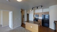 This single-floor space showcases an open-concept layout where the kitchen flows naturally into the living area. The kitchen is equipped with black appliances, including a refrigerator, electric range, and over-the-range microwave, complemented by light wood cabinetry that provides ample storage. A peninsula island with a laminate countertop serves as both a workspace and a visual divider between the kitchen and the adjoining living area. Track lighting mounted on the ceiling highlights the kitchen zone, while recessed lighting illuminates the hallway beyond. The backsplash features clean white tile, adding a bright contrast to the warm-toned cabinetry. The living area transitions from hard flooring in the kitchen to plush carpeting, creating a comfortable and distinct zone. A hallway visible from the main living space leads to additional rooms, with what appears to be a closet and utility panel access along the corridor. Neutral wall tones throughout contribute to a cohesive, understated aesthetic. The overall layout is practical and efficiently designed, making good use of the available space.