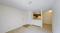 This single-floor residential space offers a straightforward and functional layout designed for comfortable everyday living. The main living area features wall-to-wall carpeting in a neutral tone, creating a soft and understated foundation for the space. White walls throughout contribute to a clean, bright atmosphere, while a textured ceiling adds a layer of subtle detail overhead.

The living area flows seamlessly toward the kitchen, which is positioned in a semi-open configuration separated by a raised pass-through counter with a light-colored surface. This design element connects the two spaces while maintaining a sense of distinct zones for cooking and relaxing. The kitchen is equipped with dark wood cabinetry that provides a warm contrast to the lighter surroundings, along with under-cabinet lighting that illuminates the workspace below. A ceiling-mounted light fixture in the kitchen ensures consistent overhead illumination.

A front entry door with a traditional six-panel design opens directly into the living area, accompanied by a closet nearby for convenient storage. A welcome mat at the entry adds a practical, homey touch. A ceiling vent is visible overhead, indicating a central HVAC system for climate control. Overall, the space presents a tidy and functional interior well-suited for residential living.