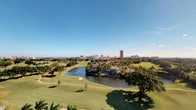 This single-floor residence offers a compelling connection to its surrounding landscape, most notably through its private balcony, which serves as a standout architectural feature. Finished in crisp white stucco with clean lines and metal railings, the balcony extends the living space outward and frames an exceptional panoramic view. From this vantage point, residents are treated to a sweeping vista of a manicured golf course, characterized by rolling fairways, mature tropical trees, and tranquil water features that wind through the grounds. In the distance, a city skyline rises above the tree line, while the horizon hints at a nearby coastline. The outdoor setting is bathed in abundant natural sunlight, reflecting the warm, clear climate of South Florida. The bright blue sky and lush greenery below create a vivid contrast against the building's white exterior. The elevated position of the balcony enhances the sense of openness and depth of the surrounding scenery. Overall, the space offers a serene residential environment defined by its access to resort-style outdoor views, tropical surroundings, and a peaceful, sun-drenched ambiance that characterizes the South Florida lifestyle.