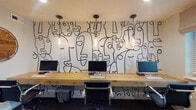 Located at 3116 Hayden Rd in Columbus, Ohio, this single-floor commercial space blends creative design with functional workspace solutions. The most visually commanding feature is a bold, large-scale abstract mural in the open work area — a continuous line-art composition of expressive, overlapping faces rendered in black and white that spans an entire wall. Beneath it, a long natural wood communal desk provides multiple workstations, each equipped with monitors, and lit from above by a row of sleek cylindrical pendant lights that lend the area a warm, modern atmosphere. The workspace strikes a balance between artistic inspiration and practical productivity. Beyond the open area, the space includes additional enclosed rooms, each featuring window blinds that allow for adjustable natural light and privacy control. These rooms offer more intimate, focused work settings suitable for individual offices, meeting spaces, or breakout areas. The overall design aesthetic is contemporary and intentional, with an emphasis on clean lines and a mix of creative and professional sensibilities. The single-floor layout ensures ease of navigation and accessibility throughout, making this Columbus workspace a compelling option for businesses seeking an environment that fosters both creativity and focused work.