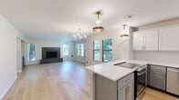 This well-appointed single-story home offers three bedrooms, two bathrooms, and a thoughtfully designed layout that maximizes both function and style. The open-concept kitchen and living area serves as the heart of the home, featuring light hardwood flooring that flows seamlessly throughout. The kitchen is outfitted with crisp white cabinetry accented by brushed gold hardware, a white quartz peninsula with an undermount sink, stainless steel appliances including a French door refrigerator, and a suite of modern pendant and flush-mount light fixtures with gold detailing. The adjacent living room is bright and welcoming, with multiple windows allowing generous natural light and views of greenery outside. A bold dark-tiled fireplace anchors the living space, adding a striking focal point. The bedrooms are carpeted for comfort, each featuring closets and windows that invite natural light. The primary bathroom offers an extended vanity with white quartz countertops, gray cabinetry with brushed hardware, a large mirror, and hardwood flooring. A second bathroom includes a tiled shower-tub combo. A dedicated laundry room houses a stacked Whirlpool washer and dryer with built-in shelving. The overall aesthetic is clean, modern, and move-in ready.