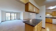 This single-floor residence offers a thoughtfully designed open-concept layout that connects the kitchen, dining, and living areas into one cohesive, flowing space. The kitchen is well-appointed with warm honey-toned wood cabinetry, dark laminate countertops, hardwood flooring, and white appliances including a range and dishwasher. A peninsula with overhead cabinetry serves as a natural divider between the kitchen and the adjoining living and dining areas, providing additional storage and counter space. The dining area is accented by an elegant chandelier, adding a refined touch to the open layout. The living area is generously proportioned with plush neutral carpet and features a sliding glass door that opens to a private balcony, inviting natural light and outdoor views into the space. The bedroom is carpeted and spacious, with its own sliding glass door providing direct balcony access. Ample closet space is present throughout the unit. The bathroom is clean and functional, featuring a tub-shower combination, a vanity with light wood cabinetry, a mirror, and tile flooring. Neutral wall tones, textured ceilings, and consistent warm wood trim details throughout lend the space a cohesive and inviting character.