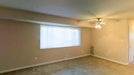 This single-floor space offers a clean, functional layout designed for comfortable everyday living. The main living area is finished with wall-to-wall carpet in a neutral tone, contributing to a warm and quiet atmosphere throughout. A large picture window fitted with vertical blinds serves as the focal point of the room, allowing an abundance of natural light to fill the space and creating an open, airy feel. A ceiling fan with an integrated light fixture is centrally positioned overhead, providing both ventilation and ambient lighting. A built-in wall unit along one side of the room offers convenient storage or display options. The walls are finished in a soft, neutral beige tone that complements the overall understated aesthetic of the space. White baseboards provide clean definition between the walls and the carpeted floor. The layout appears straightforward and versatile, lending itself well to a variety of furniture arrangements. Overall, the space presents a practical and comfortable environment with key features that support both relaxed living and everyday functionality.