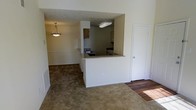 This single-floor residence offers a practical and comfortable open-concept layout that connects the main living spaces in a cohesive flow. The living area features wall-to-wall carpeting that adds warmth underfoot, transitioning to hardwood flooring near the entryway, creating a clear delineation between spaces. A centrally positioned kitchen peninsula serves as the focal point, providing a convenient pass-through design that connects the kitchen to the dining and living areas. This layout encourages easy interaction and functional use of the shared space. The kitchen is equipped with cabinetry in warm wood tones, a range, and overhead lighting, offering practical workspace within a compact footprint. The dining area is highlighted by a classic pendant light fixture that adds a touch of character to the neutral, light-toned interior. White walls and a textured ceiling reflect light throughout, keeping the space feeling bright and open. A white interior door leads to an additional room, adding to the unit's versatility. Overall, the space presents a clean, understated aesthetic with a layout designed for everyday ease and functionality.