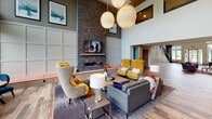 This thoughtfully designed single-floor amenity space offers a dynamic blend of comfort, functionality, and contemporary style. The centerpiece of the space is a dramatic double-height lounge area anchored by a floor-to-ceiling stone accent wall, a linear gas fireplace, and a wall-mounted television. Oversized globe pendant lights hang overhead, casting a warm glow over an eclectic mix of seating that includes bold yellow wingback chairs, a plush gray sectional, and accent tables — all arranged atop an area rug on warm wood-look flooring that runs throughout the space.

Adjacent to the lounge, floor-to-ceiling windows flood a seating alcove with natural light while offering direct views of a lush outdoor terrace complete with a pergola and patio furniture. This seamless indoor-outdoor connection enhances the airy, open ambiance of the space.

A dedicated entertainment room features a large built-in media wall with integrated LED accent lighting, ample shelving, and a generous L-shaped sectional sofa, complemented by a shuffleboard table and a patterned area rug for a social, game-room atmosphere.

The fully equipped kitchen includes dark cabinetry, stainless steel appliances, a tile backsplash, and a sleek white waterfall-edge island with seating for several, accented by teal-toned walls that add a modern, residential feel to the communal cooking and gathering area.
