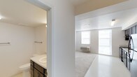 This single-floor space offers a clean and functional layout designed for comfortable everyday living. The interior is finished in a crisp, all-white color palette that enhances the sense of openness and allows natural light to flow freely throughout. The main living area is generously proportioned, featuring large windows that flood the room with natural light and create an airy, welcoming ambiance. Light-toned flooring transitions seamlessly from a tiled entry area into soft carpeting, adding warmth and comfort underfoot. A wall-mounted PTAC unit ensures climate control year-round. Adjacent to the main living space, the bathroom is thoughtfully appointed with a wide vanity topped with a white countertop and an integrated sink, complemented by dark cabinetry with brushed metal hardware that provides a subtle contrast to the otherwise bright surroundings. A towel bar and overhead lighting complete the practical yet tidy design. The overall aesthetic of the space is minimal and neutral, offering a versatile canvas that is easy to personalize. The layout is efficient and well-organized, making excellent use of the available footprint while maintaining a sense of spaciousness throughout.