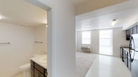 This single-floor space offers a clean and functional layout designed for comfortable everyday living. The interior is finished in a crisp, all-white color palette that enhances the sense of openness and allows natural light to flow freely throughout. The main living area is generously proportioned, featuring large windows that flood the room with natural light and create an airy, welcoming ambiance. Light-toned flooring transitions seamlessly from a tiled entry area into soft carpeting, adding warmth and comfort underfoot. A wall-mounted PTAC unit ensures climate control year-round. Adjacent to the main living space, the bathroom is thoughtfully appointed with a wide vanity topped with a white countertop and an integrated sink, complemented by dark cabinetry with brushed metal hardware that provides a subtle contrast to the otherwise bright surroundings. A towel bar and overhead lighting complete the practical yet tidy design. The overall aesthetic of the space is minimal and neutral, offering a versatile canvas that is easy to personalize. The layout is efficient and well-organized, making excellent use of the available footprint while maintaining a sense of spaciousness throughout.