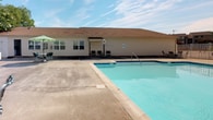 Located at 5781 Lake Edward Drive in Virginia Beach, Virginia, this single-story property offers a well-rounded combination of outdoor amenities and comfortable interior spaces. The exterior showcases a classic light-sided building with a warm brown roof, complemented by a generously sized outdoor swimming pool. The pool area is surrounded by a wide concrete deck that provides ample space for lounging, and includes patio seating with a shaded umbrella table — ideal for relaxing under the open Virginia sky.

The interior is accessed through glass-panel entry doors that open into a well-appointed hallway finished with earthy, multi-toned slate-style tile flooring. The entryway is bright and neatly maintained, with crisp white trim, paneled doors, and soft neutral wall tones that create a clean, welcoming atmosphere. Nautical-themed artwork adorns the walls, featuring framed photographs of sailboats and coastal scenes that echo the waterfront character of the Virginia Beach area. Safety features, including a fire alarm pull station, are clearly visible near the entrance.

Through the glass door, a glimpse into the adjacent interior space reveals ceiling fans, warm lighting, and additional furnishings, suggesting a comfortable common or gathering area. The overall ambiance of the property is relaxed and coastal, with thoughtful details that reflect its seaside Virginia surroundings.