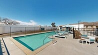 Located at 21620 Dracaea Avenue in Moreno Valley, California, this outdoor amenity area centers around a generously sized resort-style swimming pool with clear, turquoise water. The pool deck is finished with a smooth concrete surface and is equipped with multiple lounge chairs and chaise lounges, providing ample space for sunbathing and relaxation under the Southern California sun. Teal and white striped umbrellas offer shaded seating areas throughout the deck, complementing the community's clean, modern aesthetic. The pool is enclosed by a black metal security fence with a gated entry, ensuring privacy and safety for users. Tall palm trees punctuate the space, adding to the resort-like ambiance. A covered pergola structure provides additional shaded retreat space along one side of the deck. The surrounding landscape features low-maintenance desert-friendly plantings that suit the local climate. Views of the nearby mountain range and the community's contemporary multi-story residential buildings — characterized by white stucco exteriors and teal-accented windows — frame the space on multiple sides. The open-sky setting allows for abundant natural light throughout the day, making this outdoor pool area a standout communal feature of the property.
