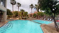 This outdoor pool area serves as a central amenity space, offering a generously sized swimming pool with clear turquoise water as its focal point. The pool is complemented by an adjacent hot tub, providing options for both active swimming and relaxing hydrotherapy. A broad concrete sun deck surrounds the pool, lined with multiple lounge chairs and umbrella stations, creating ample space for sunbathing and outdoor leisure.

The landscape design incorporates tall palm trees, mature shade trees, and native desert plantings such as agave, lending a tropical yet regionally-inspired atmosphere to the space. The surrounding multi-story residential building features brick and stucco exteriors with prominent chimneys, balconies, and pitched rooflines, forming an enclosed and private courtyard-like setting.

A dedicated pool house structure anchors one side of the deck, featuring large floor-to-ceiling windows on the upper level and a covered, gated lower area with seating. Rustic wood-framed entry doors with posted pool rules provide a clear access point to the pool area. The combination of open sky above, lush greenery, and well-maintained facilities creates an inviting and resort-like outdoor retreat for residents.