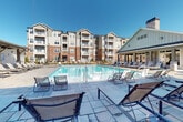 This outdoor amenity area is designed around a generously sized swimming pool set within a broad, stone-tiled sun deck lined with rows of chaise lounges. The pool features multiple entry points and reflects the open sky, creating a bright, resort-like atmosphere. Adjacent to the pool, a substantial covered pavilion serves as the centerpiece of the outdoor social space. The pavilion is constructed with a blend of board-and-batten siding, stacked stone columns, and dark wood beam detailing on the ceiling, lending a refined yet relaxed aesthetic. Inside the pavilion, a stone fireplace with a wall-mounted television anchors a comfortable seating area furnished with wicker sofas and leather cushions. Opposite the lounge area, an outdoor kitchen with a built-in grill and bar-height dining table provides a functional space for entertaining. Ceiling fans throughout the pavilion ensure comfort during warm weather. The open-air perimeter of the pavilion offers views of the surrounding residential buildings and landscaped grounds. Stone accent walls and large-format pavers tie the design elements together, creating a cohesive and polished outdoor living environment suited for relaxation and social gatherings.