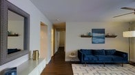 This single-floor space offers a thoughtfully designed interior that balances modern aesthetics with functional living. The living area serves as the focal point, featuring rich dark hardwood floors that contrast elegantly with the light, neutral-toned walls and ceiling, creating a clean and contemporary atmosphere.

A deep navy blue sofa anchors the seating area, complemented by a patterned area rug in soft grey and white tones that adds texture to the space. A wall-mounted floating shelf displays tasteful decorative accents, including a small vase and sculptural piece, alongside a bold blue abstract canvas that introduces a pop of color and artistic character to the room. A small round side table with a metallic gold geometric base adds a refined decorative touch nearby.

To one side, a sleek low-profile white console or storage unit hosts a gold cylindrical accent piece, reinforcing the modern design theme. The hallway beyond the living area features smooth white walls and leads to additional rooms, hinting at a well-connected and practical floor plan. Overall, the space exudes a polished, contemporary ambiance with carefully curated details throughout.