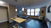 This single-floor space offers a thoughtfully designed layout that balances modern aesthetics with everyday functionality. The open living area serves as the heart of the space, featuring dark hardwood flooring, a neutral color palette, and large windows fitted with blinds that invite an abundance of natural light while offering privacy. A butcher-block prep table anchors the kitchen area, which is illuminated by a trio of pendant lights suspended from the ceiling, adding a contemporary touch. A ceiling fan provides additional comfort. The living area is comfortably arranged with a sofa, coffee table, and area rug, creating an inviting gathering space.

A hallway connects the living area to the bathroom, which is finished with a clean, bright design. The bathroom features a dual-sink vanity topped with granite countertops, white cabinetry offering ample storage, and a large mirror framed by bright overhead lighting. The hallway also includes a wall-mounted hook rack, providing a convenient spot for coats and accessories near what appears to be an entry door. Overall, the space presents a cohesive, modern aesthetic with practical details throughout.