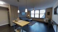 This single-floor space offers a thoughtfully designed layout that balances modern aesthetics with everyday functionality. The open living area serves as the heart of the space, featuring dark hardwood flooring, a neutral color palette, and large windows fitted with blinds that invite an abundance of natural light while offering privacy. A butcher-block prep table anchors the kitchen area, which is illuminated by a trio of pendant lights suspended from the ceiling, adding a contemporary touch. A ceiling fan provides additional comfort. The living area is comfortably arranged with a sofa, coffee table, and area rug, creating an inviting gathering space.

A hallway connects the living area to the bathroom, which is finished with a clean, bright design. The bathroom features a dual-sink vanity topped with granite countertops, white cabinetry offering ample storage, and a large mirror framed by bright overhead lighting. The hallway also includes a wall-mounted hook rack, providing a convenient spot for coats and accessories near what appears to be an entry door. Overall, the space presents a cohesive, modern aesthetic with practical details throughout.
