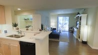 This single-floor residence showcases a thoughtfully designed open-concept layout that effortlessly connects the kitchen, dining, and living spaces into one cohesive, light-filled environment. The kitchen features a clean white breakfast bar peninsula, white cabinetry, and modern appliances including a dishwasher, making it both functional and visually appealing. The peninsula serves as a natural divider between the kitchen and the main living area, while still preserving the open flow of the space.

The living area is anchored by warm hardwood flooring that extends throughout, creating a sense of continuity and warmth. Soft, neutral wall tones in sage green and warm white contribute to a calm and welcoming ambiance. A built-in display cabinet with glass paneling adds a refined architectural detail to one corner of the room.

Natural light pours in abundantly through large sliding glass doors at the far end of the living room, which offer views of mature trees and direct access to an outdoor area. This feature not only brightens the interior but also creates a seamless indoor-outdoor connection. Recessed lighting and a floor lamp provide warm supplemental illumination throughout. Overall, the space presents a comfortable, airy, and well-appointed single-level layout ideal for everyday living.
