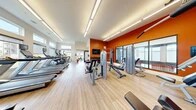 Located at 8501 Franklin Square Dr in Baltimore, MD, this fitness center offers a well-designed, single-floor space equipped to support a full range of workout routines. The interior is defined by a striking burnt-orange accent wall that energizes the space, complemented by crisp white walls and warm light wood-style flooring throughout. Recessed linear lighting runs along the ceiling, providing bright, even illumination across the entire facility.

The main workout area is generously sized, housing a comprehensive selection of strength training machines, including cable systems, weight benches, and selectorized machines arranged for easy access and flow. A dedicated dumbbell rack with free weights is mounted against the accent wall beneath a wall-mounted flat-screen television. A second, naturally lit section along the perimeter features floor-to-ceiling windows that flood the space with daylight and provide outdoor views, housing a row of treadmills, elliptical trainers, and a rowing machine. This cardio zone benefits particularly from the natural brightness, creating an open and airy atmosphere for users.

Glass doors at one end of the facility offer a transparent view into an adjacent area, enhancing the sense of openness. The overall design balances function and aesthetics, delivering an inviting and energizing environment suited for a variety of fitness activities.