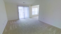 This single-floor space offers an open, airy layout characterized by neutral tones and plush wall-to-wall carpeting throughout. The main living area is generously proportioned, allowing for flexible furniture arrangements and comfortable everyday use. A sliding glass door dressed with vertical blinds serves as a prominent feature, flooding the room with natural light while providing direct access to an outdoor area. An additional window in the adjoining space further enhances the bright, welcoming atmosphere. The walls are finished in a clean, soft white, complementing the light-toned carpeting and contributing to an overall sense of openness. A subtle transition in the layout hints at a secondary space — likely a dining area or additional living zone — visible through an open passageway, suggesting a practical and connected floor plan. Ceiling-mounted HVAC vents indicate a climate-controlled environment. The uncluttered, vacant state of the space allows for a clear appreciation of its proportions, neutral palette, and abundant natural light, making it a versatile and adaptable living environment.