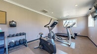 Located at 26660 E Ninth St in Highland, California, this fitness center offers a dedicated space for cardiovascular and strength training. The room is finished with neutral-toned walls, crown molding, and carpet flooring complemented by protective rubber exercise mats beneath the equipment. Recessed lighting and HVAC vents are integrated into the ceiling, ensuring a comfortable workout environment. Multiple windows line the walls, allowing natural light to fill the space and providing views of the surrounding area. The facility is equipped with a treadmill, two elliptical trainers, and a stationary exercise bike, all positioned to maximize the use of floor space. A freestanding dumbbell rack with a range of weights is available for strength training. A wall-mounted flat-screen television is installed for entertainment during workouts, and a large mirror on one wall enhances the sense of space while allowing users to monitor their form. The room is accessed through a distinctive red entry door. Overall, the fitness center presents a clean, functional, and well-organized environment suited for a variety of workout routines.