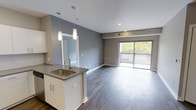 This single-floor space showcases a thoughtfully designed open-concept layout that integrates the kitchen and living areas into one cohesive environment. The kitchen features a peninsula with a stainless steel sink, sleek quartz-style countertops, and white cabinetry with contemporary hardware. Two elegant pendant lights hang above the peninsula, providing focused task lighting while adding a modern aesthetic touch. Recessed lighting throughout the ceiling ensures even, ambient illumination across the space.

Dark hardwood-style flooring runs continuously throughout, creating a rich contrast against the light gray walls and white trim. The living area is generously proportioned, offering flexible furniture arrangement options. A large sliding glass door at the far end floods the space with natural light and provides direct access to a private balcony, which overlooks a lush, tree-lined outdoor view. The combination of neutral tones, modern finishes, and abundant natural light contributes to a clean, contemporary ambiance that feels both functional and inviting.