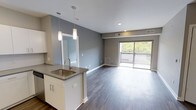 This single-floor space showcases a thoughtfully designed open-concept layout that integrates the kitchen and living areas into one cohesive environment. The kitchen features a peninsula with a stainless steel sink, sleek quartz-style countertops, and white cabinetry with contemporary hardware. Two elegant pendant lights hang above the peninsula, providing focused task lighting while adding a modern aesthetic touch. Recessed lighting throughout the ceiling ensures even, ambient illumination across the space.

Dark hardwood-style flooring runs continuously throughout, creating a rich contrast against the light gray walls and white trim. The living area is generously proportioned, offering flexible furniture arrangement options. A large sliding glass door at the far end floods the space with natural light and provides direct access to a private balcony, which overlooks a lush, tree-lined outdoor view. The combination of neutral tones, modern finishes, and abundant natural light contributes to a clean, contemporary ambiance that feels both functional and inviting.
