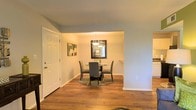 This single-floor space presents a clean, welcoming interior defined by warm hardwood flooring that extends throughout the main living areas. The dining area serves as a central feature, accommodating a round glass-top table surrounded by upholstered chairs in a muted gray tone, all anchored by a circular area rug. Above the table, a classic pendant light fixture provides focused ambient lighting, complementing the overall warm atmosphere of the room. A decorative mirror with a dark frame hangs on the wall, adding depth to the space, while a contemporary piece of wall art contributes a subtle visual accent. The walls are finished in a soft, neutral blue-gray tone, creating a calm and cohesive aesthetic throughout. White baseboards and trim provide crisp contrast against both the walls and flooring. A white paneled door near the entry adds a traditional architectural touch. The open layout between the entryway and dining zone promotes a sense of flow and accessibility, making the space feel connected and functional. Overall, the interior balances simplicity with thoughtful design details.