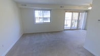 This single-floor residential space offers a clean, functional layout designed for comfortable everyday living. The main living area is generously sized, featuring wall-to-wall carpeting in a neutral tone that adds warmth underfoot. A large window along one wall floods the room with natural light, offering views of the surrounding building exterior, while a set of sliding glass doors opens directly onto a private balcony, blurring the line between indoor and outdoor living. The balcony provides an inviting outdoor retreat with views of the adjacent greenery. Neutral, light-toned walls throughout the living space create a bright and airy atmosphere, lending itself well to a variety of interior styles. Ceiling vents are positioned strategically for efficient climate control throughout the space. The overall ambiance is clean, open, and move-in ready, with a simple and practical design that maximizes the use of available space. Whether used as a primary residence or a rental unit, this space offers a welcoming environment with functional features and a calm, understated aesthetic that serves as an excellent foundation for personalization.