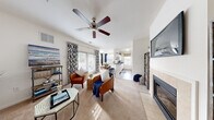 This single-floor residence offers a thoughtfully designed open-concept layout that seamlessly connects the main living spaces. The living room is a welcoming focal point, featuring plush carpeting, a ceiling fan, a tiled fireplace, and sliding glass doors that invite natural light. A wall-mounted television and built-in shelving add functional detail, while the open sightlines create a comfortable sense of spaciousness.

The adjacent dining area accommodates a round table for four beneath a large window dressed with navy drapes, providing a bright, intimate setting for meals. The kitchen is well-equipped with white cabinetry, granite-style countertops, white appliances including a range and refrigerator, and rich hardwood-style flooring. A window above the counter furthers the light-filled atmosphere.

The bedrooms are carpeted and comfortably sized. One bedroom features twin beds, wall art, and a pendant light fixture, while another offers space for a single bed and a study area. A spacious walk-in closet with multi-tier wire shelving provides generous storage. The full bathroom includes a wide vanity with ample counter space, a large mirror, white cabinetry, tile flooring, and a shower-tub combination.