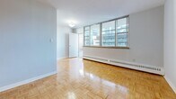 This well-appointed 2-bedroom, 1-bathroom residence is laid out across a single floor, offering a practical and comfortable living arrangement. Throughout the main living areas and bedrooms, warm-toned parquet hardwood flooring adds a timeless, classic character to the space. The soft blue-grey wall tones create a calm, neutral backdrop that complements the natural light filtering in through generously sized windows.

The living room is a bright, open area featuring large windows that frame views of the surrounding urban cityscape and allow abundant natural light to fill the room. Baseboard heating runs along the perimeter, ensuring year-round comfort. The primary bedroom is a well-proportioned retreat complete with mirrored sliding closet doors and a window with outdoor views. The second bedroom is similarly finished, with its own window and the same gleaming parquet flooring.

The galley-style kitchen is efficiently designed with white cabinetry fitted with brass hardware, granite-look countertops, a stainless steel sink, and white appliances including a gas range and full-sized refrigerator. Classic tile flooring with a decorative pattern adds a charming detail. The bathroom features a full soaking tub with white subway tile surround. An entrance and hallway connect all areas of the home seamlessly.