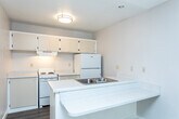Located at 1080 W 3300 S in South Salt Lake, Utah, this efficient studio unit offers a well-organized single-floor layout that combines a kitchen and living area with one full bathroom. The open-concept kitchen is equipped with white appliances, including an electric range, refrigerator, and a range hood, complemented by cream-toned upper cabinetry and a light-speckled countertop peninsula that doubles as a breakfast bar with an integrated double-basin sink. The peninsula design creates a natural division between the kitchen and the living area, maximizing the use of the compact footprint. Wood-look laminate flooring runs throughout the space, adding warmth and visual continuity. The living area benefits from a large window fitted with vertical blinds, allowing generous natural light to filter in while offering views of the surrounding outdoor greenery. A wall-mounted HVAC unit provides climate control. The bathroom features white tile surrounding a full bathtub and shower combination, maintaining a clean and functional aesthetic. Neutral wall tones and a textured ceiling in the living area contribute to a simple, understated ambiance throughout this practical and self-contained unit.