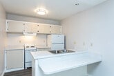 Located at 1080 W 3300 S in South Salt Lake, Utah, this efficient studio unit offers a well-organized single-floor layout that combines a kitchen and living area with one full bathroom. The open-concept kitchen is equipped with white appliances, including an electric range, refrigerator, and a range hood, complemented by cream-toned upper cabinetry and a light-speckled countertop peninsula that doubles as a breakfast bar with an integrated double-basin sink. The peninsula design creates a natural division between the kitchen and the living area, maximizing the use of the compact footprint. Wood-look laminate flooring runs throughout the space, adding warmth and visual continuity. The living area benefits from a large window fitted with vertical blinds, allowing generous natural light to filter in while offering views of the surrounding outdoor greenery. A wall-mounted HVAC unit provides climate control. The bathroom features white tile surrounding a full bathtub and shower combination, maintaining a clean and functional aesthetic. Neutral wall tones and a textured ceiling in the living area contribute to a simple, understated ambiance throughout this practical and self-contained unit.