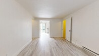This single-floor unit offers a clean, minimalist aesthetic throughout, characterized by crisp white walls, textured ceilings, and light wood-style luxury vinyl flooring that runs consistently across the living spaces. The interior is organized into multiple rooms, each finished with wide white baseboards and a neutral color palette that creates a bright, cohesive feel.

The kitchen is outfitted with white cabinetry, stainless steel appliances including a refrigerator, electric range, microwave, and dishwasher, along with durable countertops and a sink. Adjacent to the kitchen, a dining or living area benefits from generous natural light provided by a sliding glass door that opens directly to a private outdoor patio enclosed by a wooden fence. A ceiling fan overhead adds to the room's functionality.

The remaining rooms are open and versatile, each featuring baseboard heating units to ensure year-round comfort. A wall-mounted air conditioning unit is also present in one room, supplementing climate control options. The bathroom includes a full soaking tub with a shower, surrounded by clean, large-format white tile and polished chrome fixtures. Throughout the unit, the overall ambiance is light, airy, and move-in ready.