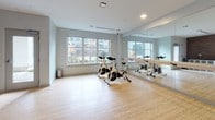 Located at 200 Presidential in Woburn, Massachusetts, this single-floor fitness studio is thoughtfully designed to support a range of exercise activities. The space is divided into distinct functional zones that flow seamlessly into one another.

The cycling area is a standout feature, equipped with professional Spinning bikes arranged for group classes. Large windows along one wall flood the room with natural light and provide views of the surrounding exterior, while a full-length wall mirror with a mounted ballet barre amplifies the sense of space and supports form-checking during workouts. The light bamboo-style flooring throughout adds warmth and is well-suited for high-activity use.

Adjacent to the cycling zone is a versatile open studio area, ideal for yoga, stretching, or group fitness classes. This room also benefits from ample natural light and mirrors, creating an open, airy atmosphere. The same clean flooring carries through, maintaining visual continuity.

A prominent accent wall clad in dark wood paneling serves as a striking focal point, anchoring a built-in media console with a large wall-mounted flat-screen television. Beneath the screen, generous white cabinetry and open shelving provide ample storage for equipment and accessories.

The overall design palette — soft gray walls, bright recessed lighting, and natural wood tones — creates a clean, modern, and motivating environment well-suited for an active lifestyle.