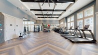 Located at Avida Village Circle in Orlando, FL, this expansive fitness center offers a generously sized, single-floor layout designed to accommodate a full range of workout activities. The space makes an immediate impression with its soaring coffered ceilings accented by dark exposed beams and large industrial ceiling fans, creating an airy, open atmosphere throughout.

The striking herringbone-patterned hardwood flooring runs the entire length of the facility, lending both elegance and a sense of continuity to the space. Rows of large windows along multiple walls flood the interior with abundant natural light while offering views of the surrounding outdoor environment, contributing to an uplifting and motivating workout environment.

The fitness center is comprehensively equipped across dedicated zones. A cardio area lines one side of the room with a full row of treadmills, ellipticals, stationary bikes, and rowing machines. The strength training area features cable machines, a power rack with a barbell and weight plates, a dumbbell rack with benches, and a variety of functional training tools including kettlebells, medicine balls, and balance equipment organized on a tiered rack. Full-length mirrors throughout the space enhance the sense of openness and allow for form monitoring during exercise.

A dedicated entry vestibule with glass double doors features an eye-catching built-in rock climbing wall, adding a unique and dynamic element to the facility.