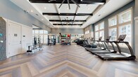 Located at Avida Village Circle in Orlando, FL, this expansive fitness center offers a generously sized, single-floor layout designed to accommodate a full range of workout activities. The space makes an immediate impression with its soaring coffered ceilings accented by dark exposed beams and large industrial ceiling fans, creating an airy, open atmosphere throughout.

The striking herringbone-patterned hardwood flooring runs the entire length of the facility, lending both elegance and a sense of continuity to the space. Rows of large windows along multiple walls flood the interior with abundant natural light while offering views of the surrounding outdoor environment, contributing to an uplifting and motivating workout environment.

The fitness center is comprehensively equipped across dedicated zones. A cardio area lines one side of the room with a full row of treadmills, ellipticals, stationary bikes, and rowing machines. The strength training area features cable machines, a power rack with a barbell and weight plates, a dumbbell rack with benches, and a variety of functional training tools including kettlebells, medicine balls, and balance equipment organized on a tiered rack. Full-length mirrors throughout the space enhance the sense of openness and allow for form monitoring during exercise.

A dedicated entry vestibule with glass double doors features an eye-catching built-in rock climbing wall, adding a unique and dynamic element to the facility.
