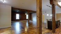 This single-floor space embodies classic loft-style architecture, seamlessly blending industrial character with open, airy living. The expansive main area is anchored by original wide-plank hardwood floors that reflect natural light, lending warmth to the overall aesthetic. Prominent structural timber columns run from floor to ceiling, serving as defining architectural features that highlight the building's historic construction. Exposed brick walls line the perimeter, adding rich texture and a sense of urban authenticity throughout the space. The high ceilings, supported by visible wooden beams, amplify the sense of volume and openness. Multiple windows allow natural light to filter in, brightening the interior while framing views of the outdoors. Transitioning toward one end of the space, a partially visible kitchen area introduces modern elements, including a countertop and pendant lighting, creating a subtle contrast against the raw, industrial backdrop. The open floor plan offers flexibility for a variety of layouts and uses. Overall, the space strikes a compelling balance between historic architectural detail and functional, contemporary living.