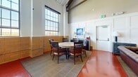 Located in Lorton, Virginia, this expansive single-floor gathering space offers a striking blend of industrial architecture and refined interior design. The most commanding feature is the dramatically vaulted ceiling with exposed dark wood trusses, structural beams, and visible ductwork, all of which lend an airy, loft-like character to the interior. Polished terracotta-red concrete floors run throughout, adding warmth and continuity across the space.

The main lounge area is anchored by rich, tufted brown leather Chesterfield sofas and accent chairs arranged around a central coffee ottoman upholstered in bold red. Crimson velvet armchairs and additional seating clusters provide flexible arrangements for socializing or events. Wainscoted white panel walls serve as an elegant backdrop, adorned with framed artwork throughout.

A full kitchenette and bar area features dark wood cabinetry, open shelving, a stainless steel refrigerator, and counter seating with industrial-style barstools. A mounted flat-screen television anchors this section of the space. A separate dining area accommodates a round table with leather-upholstered chairs beneath tall multi-pane windows that draw in natural light. The combination of soaring ceilings, curated furnishings, and versatile layout makes this a highly functional and visually distinctive venue.