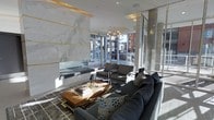 This single-floor space showcases a refined contemporary design with an emphasis on high-end materials and elegant detailing. The focal point of the living area is a dramatic floor-to-ceiling marble feature wall adorned with bold gold inlay accents, housing a sleek glass-enclosed fireplace that adds both warmth and visual impact. The white and grey veining of the marble creates a striking contrast against the gold elements, contributing to the space's upscale aesthetic.

Large floor-to-ceiling windows line the perimeter, drawing in abundant natural light and providing open views of the surrounding streetscape. The transparency of the glazing creates a seamless connection between the interior and the urban environment outside. A cylindrical stone or concrete column near the window line adds an architectural punctuation to the open layout.

The lounge area is furnished with plush charcoal grey sofas, complemented by patterned accent cushions and a warm wood-and-metal coffee table with lucite detailing. A pendant chandelier overhead adds a decorative touch. The overall ambiance is polished, modern, and inviting, balancing luxury materials with comfortable, livable furnishings.