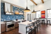 This single-floor communal space combines contemporary design with warm, inviting elements to create a highly functional and visually striking environment. The open-plan layout is anchored by soaring ceilings adorned with exposed dark wood beams, which contrast beautifully against crisp white walls and rich hardwood flooring that runs throughout the space.

The kitchen area is a standout feature, showcasing a bold blue hexagonal tile backsplash with decorative snowflake detailing, wood-tone cabinetry, white countertops, and a professional stainless steel range with a sleek hood. A central island with seating provides an informal gathering point, complemented by statement pendant lighting in warm gold tones.

Adjacent to the kitchen, a high-top dining area offers multiple bar-height tables and black metal stools, bathed in natural light from large picture windows that frame views of the surrounding exterior. Bold abstract artwork in warm orange and teal tones accents the walls, adding color and personality to the space.

The lounge area features a generous sectional sofa, a swivel accent chair, a wall-mounted television, and a glass coffee table, all arranged on a layered area rug. Large windows on multiple walls flood this seating zone with natural light and provide open views of the neighboring outdoor areas, creating a bright and connected atmosphere throughout.