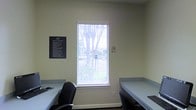 This single-floor workspace is designed with functionality and efficiency in mind. The room features two built-in workstations positioned along opposing walls, each offering ample desk surface for productive use. The desks have a sleek, utilitarian finish in a muted gray-blue tone, complementing the neutral, light-colored walls that contribute to a clean and professional atmosphere.

A centrally placed window fitted with white horizontal blinds serves as the focal point of the room, allowing natural light to filter in while maintaining privacy. The view through the window reveals mature trees and greenery, offering a subtle connection to the outdoors. The window's placement between the two workstations ensures both users benefit equally from the natural light.

The space is equipped with ergonomic seating, including a black executive-style chair, supporting comfort during extended work sessions. Informational signage is mounted on the walls, including a posted rules or guidelines board and a no-smoking notice, indicating this is a shared or managed workspace. The overall ambiance is calm, organized, and purposeful, making it well-suited for focused individual or collaborative work.