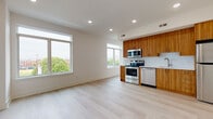 This well-designed single-floor unit offers a clean, modern aesthetic with light hardwood flooring and crisp white walls throughout. The open-concept living and kitchen area is the heart of the space, bathed in natural light from oversized windows that frame outdoor tree and cityscape views. The kitchen is thoughtfully appointed with rich wood-grain cabinetry extending to the ceiling, a white tile backsplash, light quartz countertops, and a full suite of stainless steel appliances including a range, microwave, dishwasher, and refrigerator. Recessed lighting illuminates the entire open living area, adding to the bright and airy atmosphere. The bedroom is generously proportioned with its own large window, a ceiling fan, and dual closets providing ample storage. The full bathroom is outfitted with floor-to-ceiling white subway tile, a soaking tub with shower, a wall-mounted vanity with an integrated sink, a large mirror, and neutral porcelain tile flooring. A dedicated entry area with recessed lighting and a secure door completes the layout. The unit's consistent use of light tones, quality materials, and abundant natural light creates a cohesive and inviting living environment.