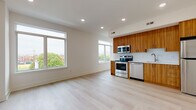 This well-designed single-floor unit offers a clean, modern aesthetic with light hardwood flooring and crisp white walls throughout. The open-concept living and kitchen area is the heart of the space, bathed in natural light from oversized windows that frame outdoor tree and cityscape views. The kitchen is thoughtfully appointed with rich wood-grain cabinetry extending to the ceiling, a white tile backsplash, light quartz countertops, and a full suite of stainless steel appliances including a range, microwave, dishwasher, and refrigerator. Recessed lighting illuminates the entire open living area, adding to the bright and airy atmosphere. The bedroom is generously proportioned with its own large window, a ceiling fan, and dual closets providing ample storage. The full bathroom is outfitted with floor-to-ceiling white subway tile, a soaking tub with shower, a wall-mounted vanity with an integrated sink, a large mirror, and neutral porcelain tile flooring. A dedicated entry area with recessed lighting and a secure door completes the layout. The unit's consistent use of light tones, quality materials, and abundant natural light creates a cohesive and inviting living environment.