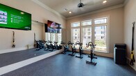 This single-floor fitness studio is designed to support both cycling-focused and functional training workouts. The spin cycling area is the centerpiece of the space, lined with a row of Matrix stationary bikes arranged in front of a full-length wall mirror that amplifies the sense of depth and allows riders to monitor their form. Large, multi-pane windows line one wall, flooding the room with natural light and offering views of lush greenery and surrounding outdoor spaces. Wall-mounted flat-screen televisions provide visual entertainment during sessions, while a ceiling fan overhead helps maintain airflow and comfort. The flooring throughout is a durable, cushioned rubber-style surface well-suited for high-intensity use.

A secondary functional training area connects to the cycling space, accessible through glass-paneled doors. This zone is outfitted with TRX suspension trainers mounted to the wall, a rack of medicine balls and resistance tools, and a collection of stability and exercise balls in various sizes. Step platforms and additional accessories are also stored here, making the area versatile for group or individual workouts. Crown molding and neutral wall tones lend the studio a refined, polished aesthetic that balances its athletic function with an inviting atmosphere.