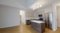 This two-floor space showcases a thoughtfully designed interior with a clean, modern aesthetic. The kitchen serves as a central highlight, featuring stainless steel appliances, including a refrigerator, gas range, built-in microwave, and dishwasher. Light grey upper cabinetry contrasts elegantly with the dark wood-toned lower cabinets and island base, while white quartz countertops provide a crisp, polished finish. The central kitchen island offers additional workspace and a natural gathering point within the space. Light hardwood flooring runs continuously throughout, contributing warmth and cohesion to the open layout. A tray ceiling detail above the kitchen adds architectural depth, complemented by a modern track lighting fixture that ensures ample task illumination. The open sightlines connect the kitchen to adjacent areas, enhancing the sense of spaciousness. Neutral white walls throughout maintain a bright, airy ambiance. A hallway visible in the background hints at additional rooms beyond, including what appears to be a laundry area, adding to the functional versatility of this well-appointed two-story space.
