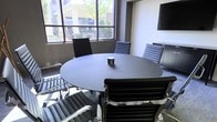 This single-floor conference room is thoughtfully designed to facilitate focused meetings and collaborative discussions. At the heart of the space sits a sleek, round dark-topped table surrounded by ergonomic high-back chairs upholstered in dark fabric, providing a professional and comfortable setting for small group meetings. A built-in power module integrated into the tabletop ensures convenient access to electrical and connectivity options for attendees.

Natural light flows generously into the room through large, multi-pane windows that line one corner of the space, offering views of a lush, green exterior environment. This abundance of natural light creates a bright and inviting atmosphere, helping to keep the room feeling open despite its intimate scale.

A wall-mounted flat-screen display is positioned prominently for easy visibility from all seats, supporting presentations and video conferencing needs. The walls are finished in a neutral gray tone, lending a modern and professional aesthetic that complements the dark furnishings. Low-pile carpeting in a muted tone adds acoustic comfort underfoot. A storage credenza is positioned against one wall, providing additional functionality. Overall, this conference room balances practicality with a clean, contemporary design suited for productive professional interactions.