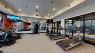 This well-appointed fitness center at 3140 E Michigan Ave in Lansing, Michigan, offers a comprehensive workout environment designed for a wide range of exercise routines. The space is finished with a neutral palette accented by bold orange walls and vibrant striped carpeting, creating an energetic yet functional atmosphere throughout.

The cardio zone features a row of Life Fitness treadmills and a selection of elliptical and cycling machines, all positioned to take advantage of the natural light filtering through large windows with horizontal blinds. Large wall-mounted mirrors amplify the sense of space and allow users to monitor their form during workouts.

A central cable resistance machine serves as the anchor of the strength training area, supplemented by a dedicated dumbbell rack offering a range of free weights. Additional functional training accessories include stability balls, resistance bands, exercise mats, and medicine balls, all neatly organized on a freestanding rack for easy access.

Mounted flat-screen televisions are positioned throughout the space for entertainment during workouts. Recessed lighting and a ceiling fan ensure the environment remains bright and comfortable. Large glass doors connect the fitness center to an adjacent amenity area, enhancing the open, community-oriented feel of the facility.