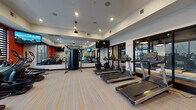 This well-appointed fitness center at 3140 E Michigan Ave in Lansing, Michigan, offers a comprehensive workout environment designed for a wide range of exercise routines. The space is finished with a neutral palette accented by bold orange walls and vibrant striped carpeting, creating an energetic yet functional atmosphere throughout.

The cardio zone features a row of Life Fitness treadmills and a selection of elliptical and cycling machines, all positioned to take advantage of the natural light filtering through large windows with horizontal blinds. Large wall-mounted mirrors amplify the sense of space and allow users to monitor their form during workouts.

A central cable resistance machine serves as the anchor of the strength training area, supplemented by a dedicated dumbbell rack offering a range of free weights. Additional functional training accessories include stability balls, resistance bands, exercise mats, and medicine balls, all neatly organized on a freestanding rack for easy access.

Mounted flat-screen televisions are positioned throughout the space for entertainment during workouts. Recessed lighting and a ceiling fan ensure the environment remains bright and comfortable. Large glass doors connect the fitness center to an adjacent amenity area, enhancing the open, community-oriented feel of the facility.