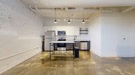Located at 612 W Main Ave in Gastonia, NC, this single-floor unit showcases a compelling blend of industrial character and modern finishes. Polished concrete floors flow throughout the entire space, reflecting light and reinforcing the open, loft-inspired aesthetic. The ceilings are a standout feature, with exposed beams, painted white planks, and visible ductwork that lend an authentic industrial charm, while pendant lighting adds warmth and definition to key areas.

The kitchen is thoughtfully designed with white upper cabinetry, gray lower cabinets, and light quartz countertops. A full suite of stainless steel appliances — including a refrigerator, range, microwave, and dishwasher — makes it highly functional. Rustic open shelving on pipe brackets adds a stylish, utilitarian touch. A painted white brick accent wall anchors the kitchen area, adding texture and depth to the overall design.

The living area is generously proportioned, offering flexible use of space, while the unit also includes a spacious walk-in closet outfitted with wire shelving systems for ample storage. A stacked washer and dryer unit is conveniently integrated within the living space. Clean white walls, modern black door hardware, and a crisp, minimalist hallway complete the cohesive industrial-modern aesthetic throughout.