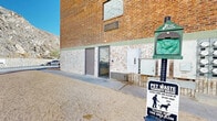This property presents a striking visual contrast between its classic multi-story brick construction and the dramatic natural rock cliffs that rise directly behind it. The building features warm red brick façades with lighter brick detailing along the base, green-trimmed windows, and a well-maintained exterior that spans multiple floors.

The surrounding grounds are equally notable. A generously sized paved parking lot wraps around the building, bordered by carefully constructed stone retaining walls that follow the natural contour of the rocky terrain. These walls serve both a functional and aesthetic purpose, integrating the built environment with the rugged desert landscape. Red curbing delineates the parking areas clearly throughout.

The property is pet-friendly, as evidenced by a dedicated pet waste station prominently positioned near one of the building's entry points. Multiple exterior entry doors provide convenient access, and a wall-mounted mailbox cluster is visible on the building's exterior, indicating a multi-unit structure.

Infrastructure components, including utility boxes and a fire hydrant, are thoughtfully positioned around the grounds. Desert-adapted landscaping with gravel ground cover and sparse native shrubs complements the arid environment. The clear blue skies and sun-drenched setting highlight the property's remarkable backdrop — a towering, striated rock face that lends the site an unmistakable and dramatic sense of place.
