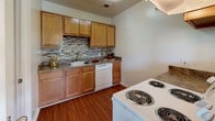 This single-floor residence offers a practical and comfortable layout with several distinct living areas. The kitchen is a functional highlight, featuring warm honey-toned wood cabinetry, granite-style countertops, and a decorative tile backsplash with a mosaic pattern. White appliances, including a coil-burner range and dishwasher, complement the rich hardwood-style flooring that runs throughout the kitchen and bathroom areas.

The open living area is bright and spacious, finished with neutral beige carpet and light gray walls. A chandelier provides an elegant focal point, while a pass-through opening connects the living space to the kitchen, enhancing the sense of openness. Multiple doorways lead to the bedrooms and additional areas of the unit.

The bedrooms are carpeted in a neutral tone and feature light gray walls, textured ceilings, and windows with blinds that allow natural light and exterior views of surrounding greenery. Each room offers a clean, versatile space suitable for various furniture arrangements.

The bathroom includes hardwood-style flooring, a vanity with warm wood cabinetry, and a full tub-shower combination with a curtain rod. The overall ambiance of the space is neutral and move-in ready, with consistent finishes throughout.