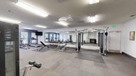 Located at 196 Jeff Rd NW in Huntsville, Alabama, this fitness center offers a comprehensive workout facility spread across a single floor with two dedicated exercise rooms. The space is designed with functionality and comfort in mind, featuring neutral-toned carpet flooring complemented by protective rubber mat sections beneath the equipment.

The main exercise room is generously sized, bathed in bright fluorescent overhead lighting that ensures a well-lit environment throughout. A large wall-mounted mirror spans a significant portion of one wall, enhancing the sense of openness and allowing users to monitor their form during workouts. A wall-mounted flat-screen television adds convenience for cardio sessions.

Equipment is thoughtfully arranged throughout the space and includes multiple treadmills, an elliptical trainer, a stationary bike, a cable machine, a leg press machine, a weight bench, and a dumbbell rack stocked with a full range of free weights and medicine balls. A secondary exercise room provides additional strength training stations and cable equipment.

Natural light filters in through several windows fitted with white blinds, some of which offer views of an outdoor pool area. Accent walls in a deep navy blue provide a modern, energizing aesthetic against the predominantly white walls. The facility also includes ceiling fans and safety amenities such as a fire extinguisher, making it a practical and inviting space for a complete fitness routine.